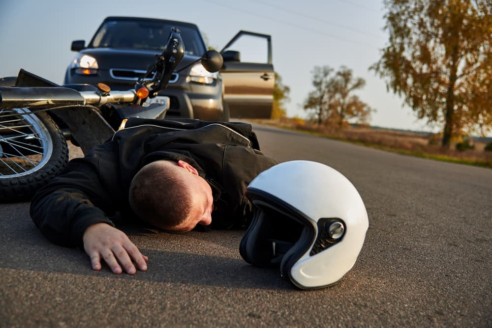 A motorcyclist is sprawled on the asphalt beside a motorcycle and car, emphasizing the aftermath of a road accident and its serious implications.