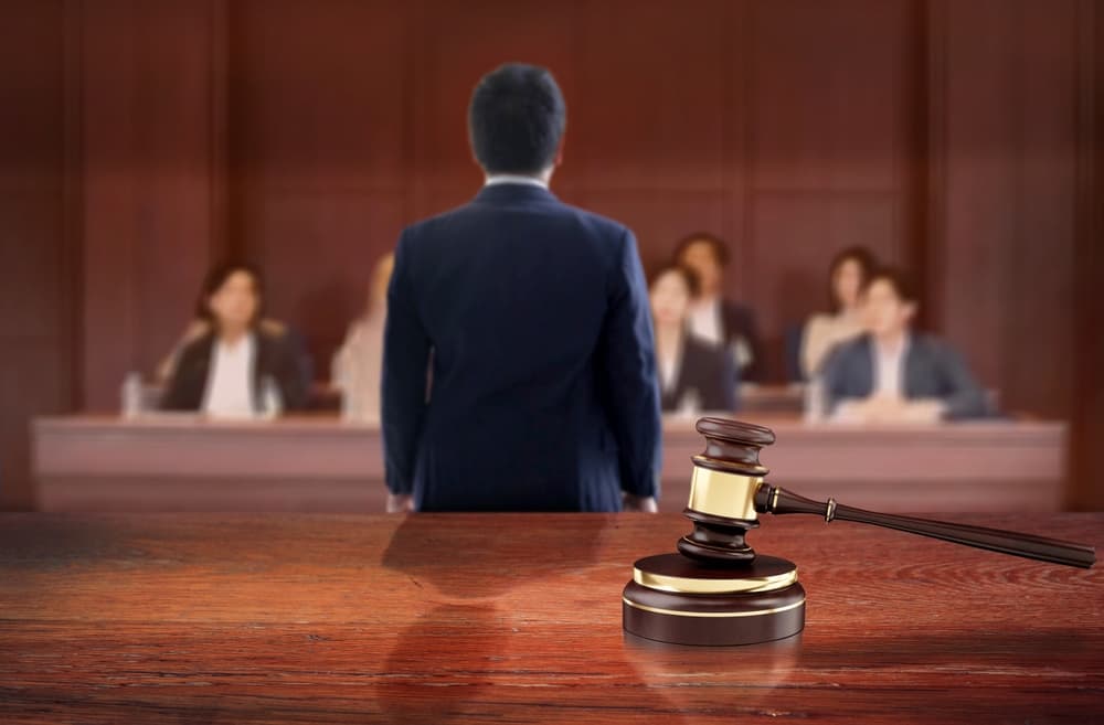 A judge's gavel symbolizes justice on a wooden courtroom table, while a lawyer addresses the jury in the background.