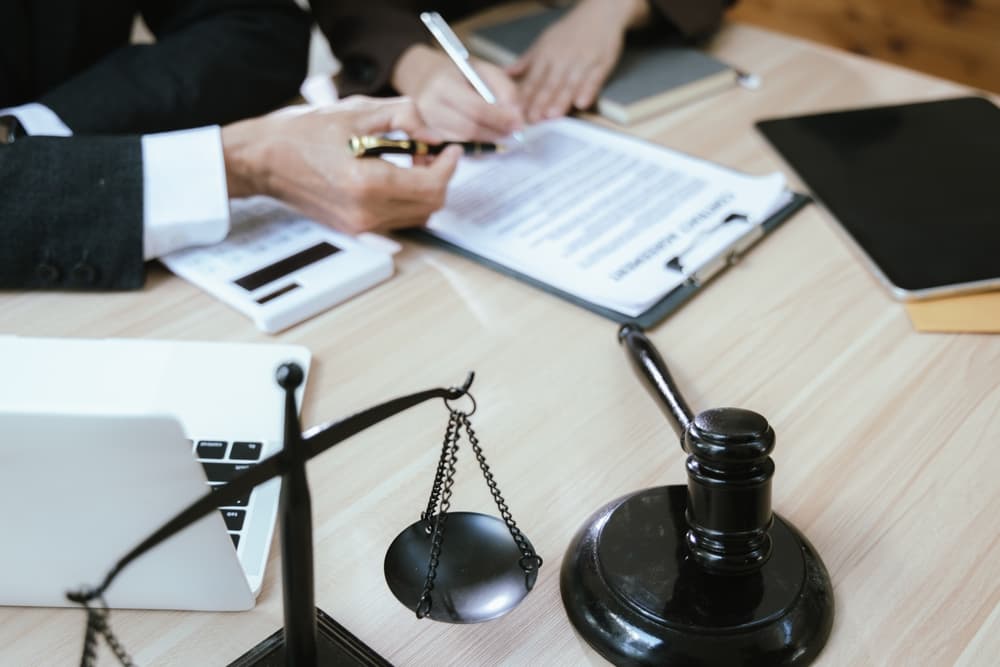 A close-up of a male lawyer in a suit working with contract documents and a wooden gavel on a courtroom table, symbolizing justice and law.