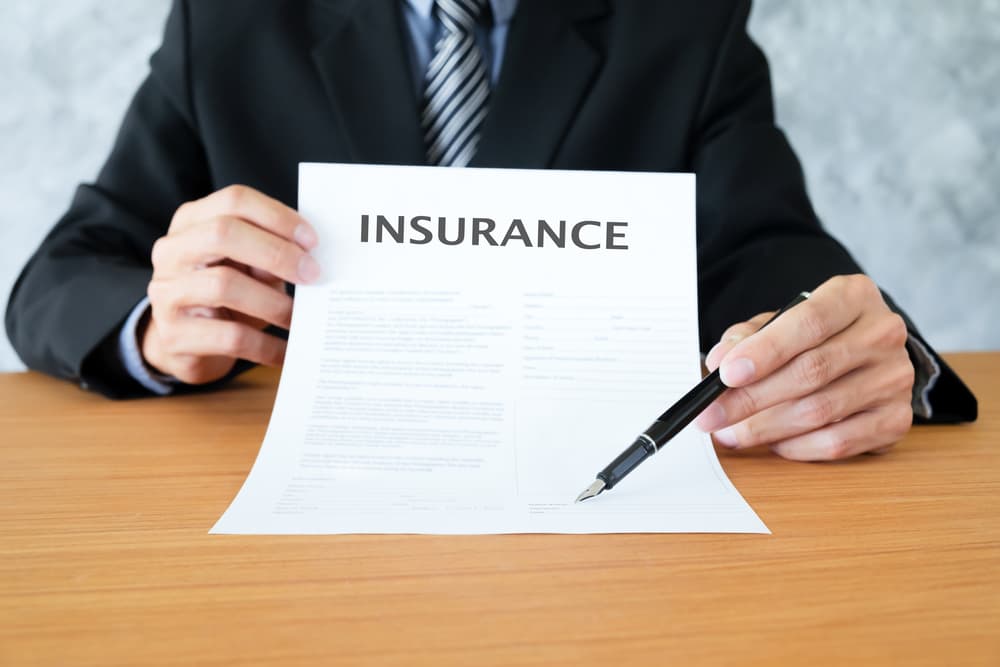 A young businessman in a suit sits in his office, holding an insurance policy and pointing with a pen to the section where the policyholder needs to sign.







